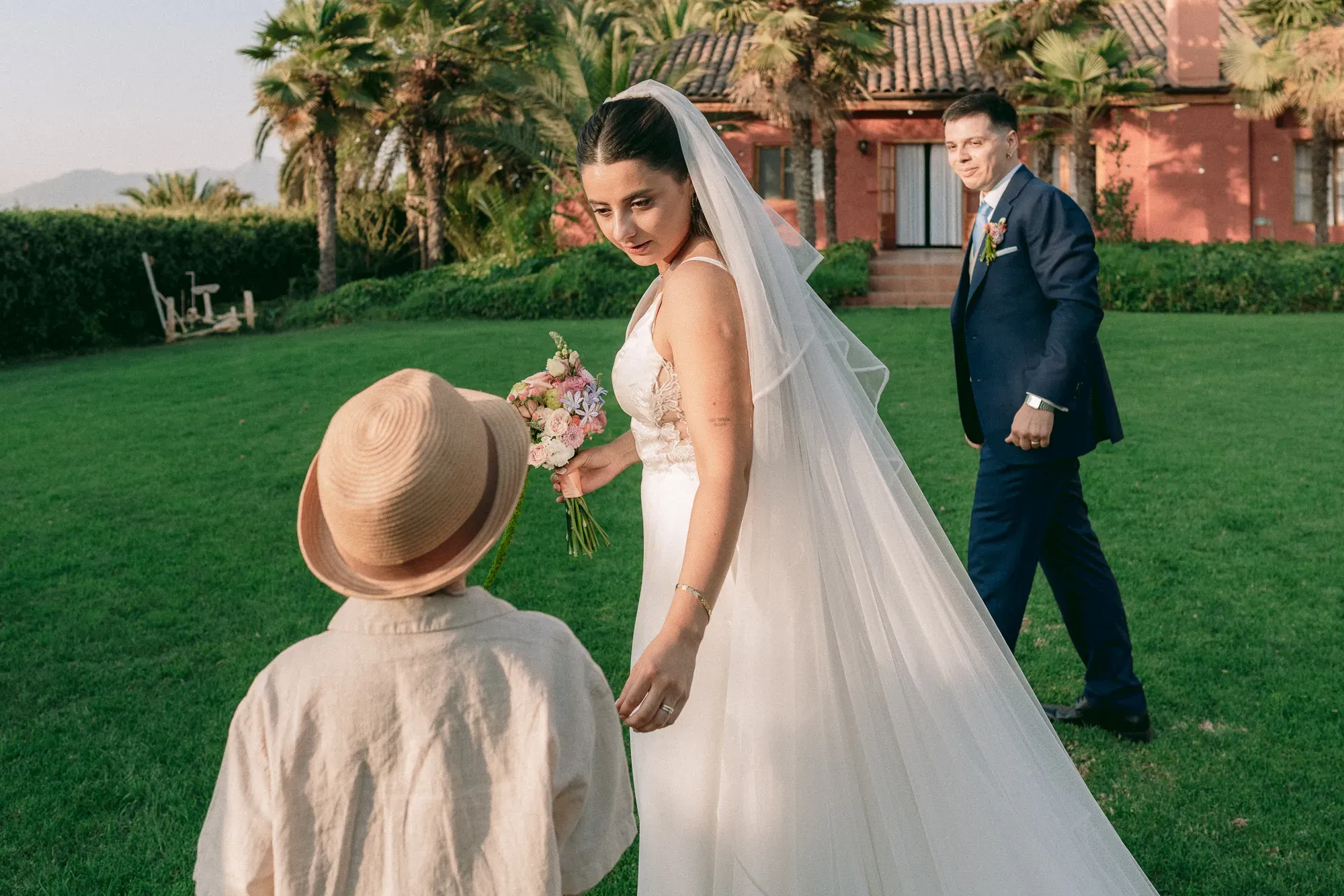 La novia mira a un niño con sombrero mientras el novio camina por el pasto