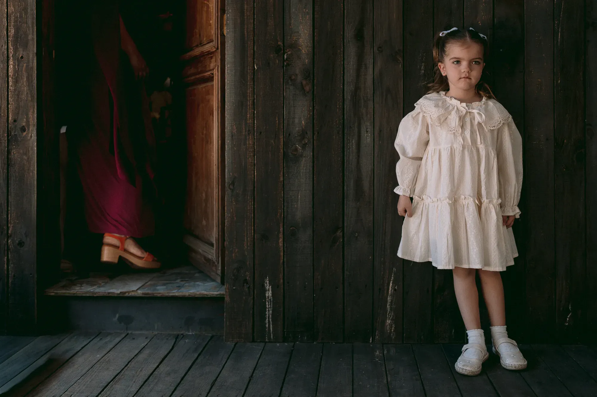 Una niña de vestido blanco, seria, contra una pared de madera