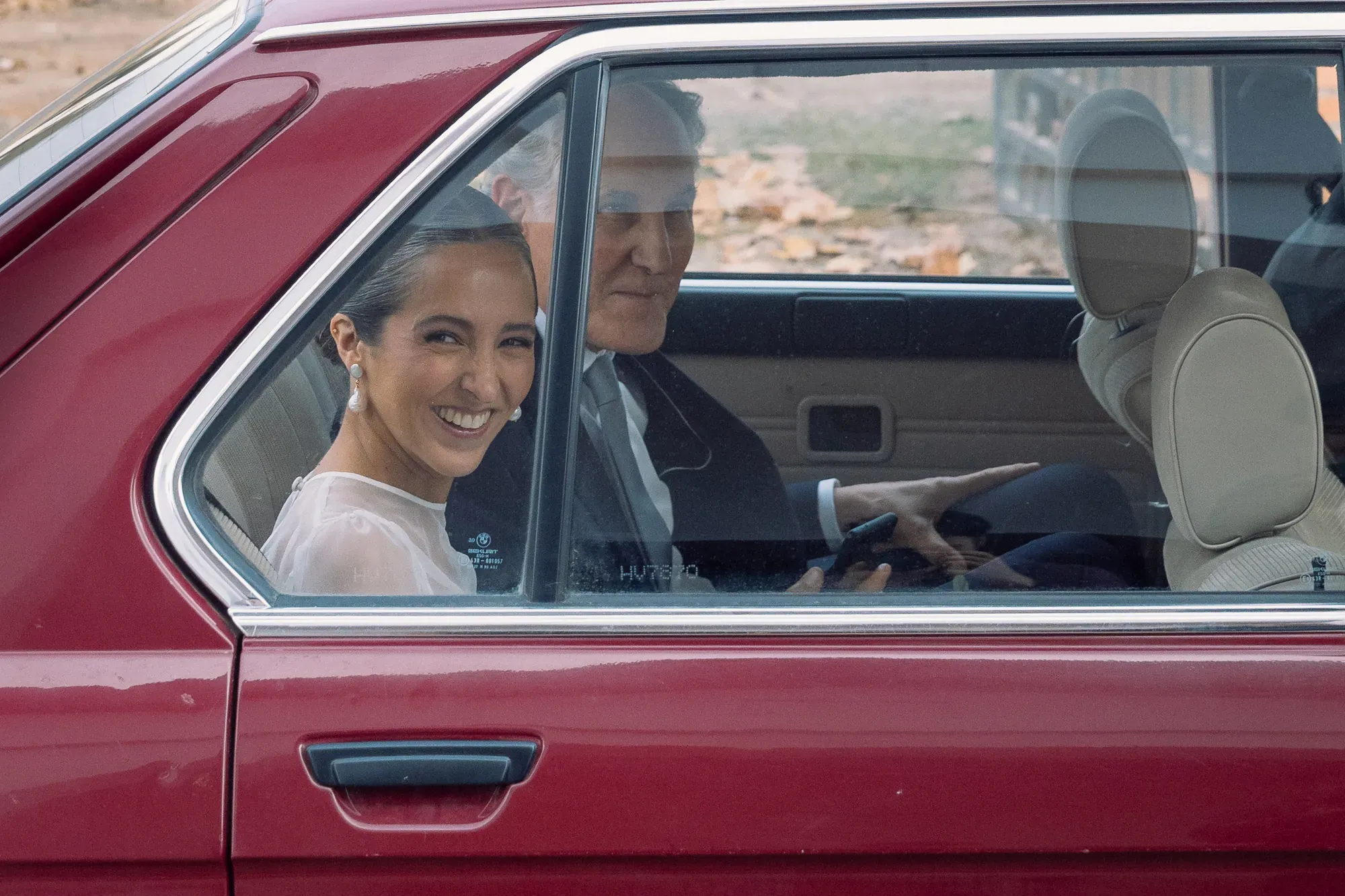 La novia sonríe desde un auto rojo antiguo